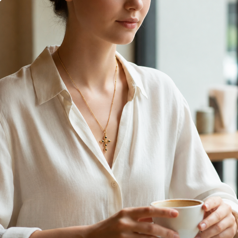 A woman in a cream-colored linen shirt holds a white ceramic cup while wearing a delicate gold cross pendant necklace with black onyx inlay, made by Sowell Jewelry.