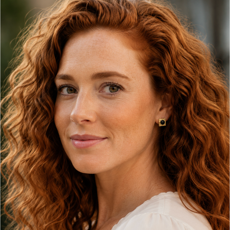 A close-up portrait of a woman with long, curly red hair wearing a white top and a small gold earring with a black stone centered, designed by Sowell Jewelry.