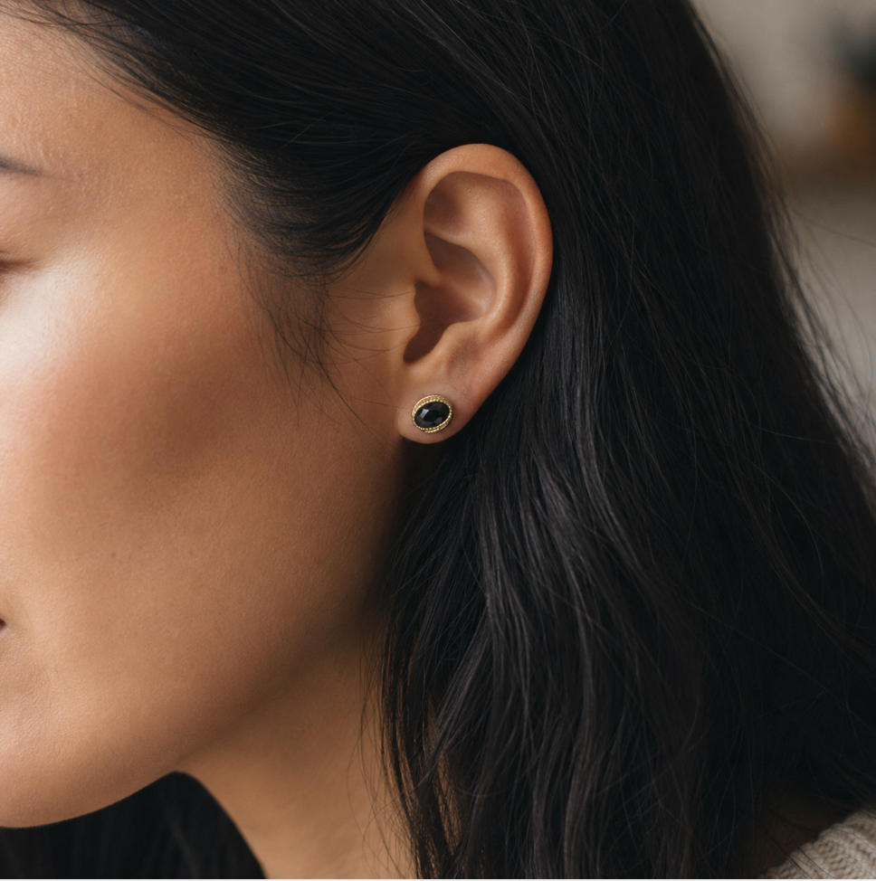 Close-up of a woman's ear wearing a small round stud earring with a dark stone set in a gold-tone bezel, designed by Sowell Jewelry.