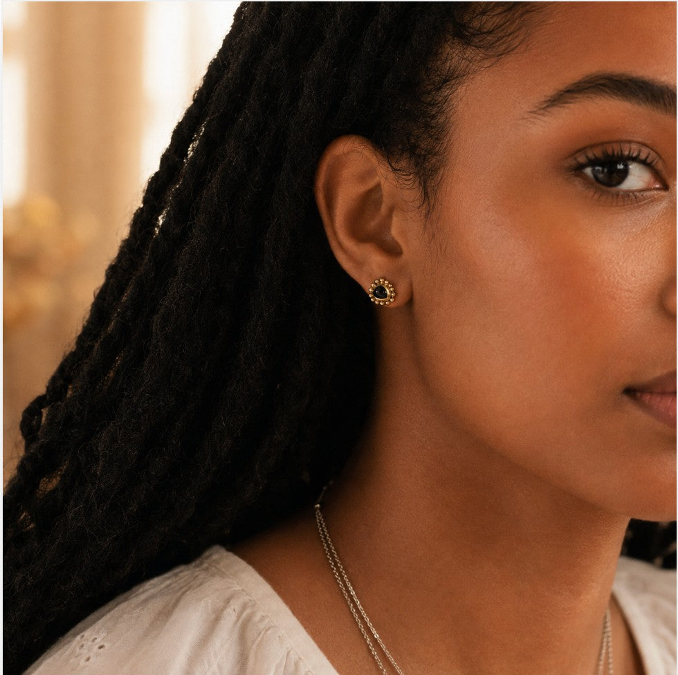A close-up lifestyle shot of a woman with dark dreadlocks wearing a small gold earring with a black stone center, designed by Sowell Jewelry.