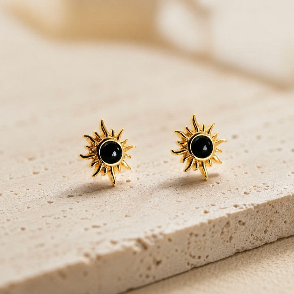 SOWELL JEWELRY 18K gold sunburst stud earrings featuring a central genuine black onyx stone on a textured stone surface.