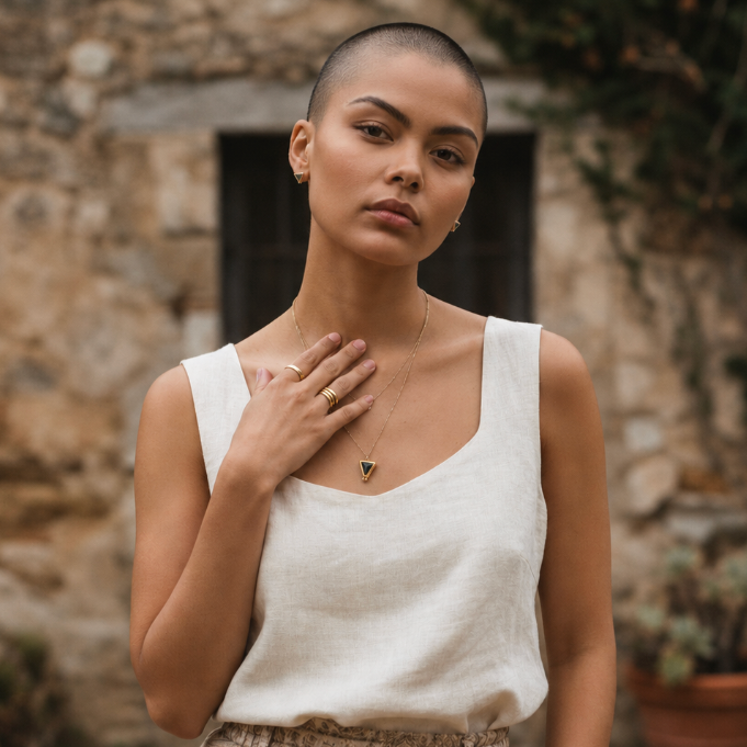 A woman with a shaved head stands outdoors in front of a rustic stone wall, wearing a delicate gold necklace with a small black stone triangular pendant and matching earrings designed by Sowell Jewelry.
